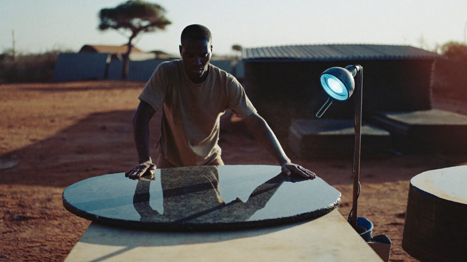 Granite fabricator in a Gweru yard, late dry-season sun raking a polished black slab.