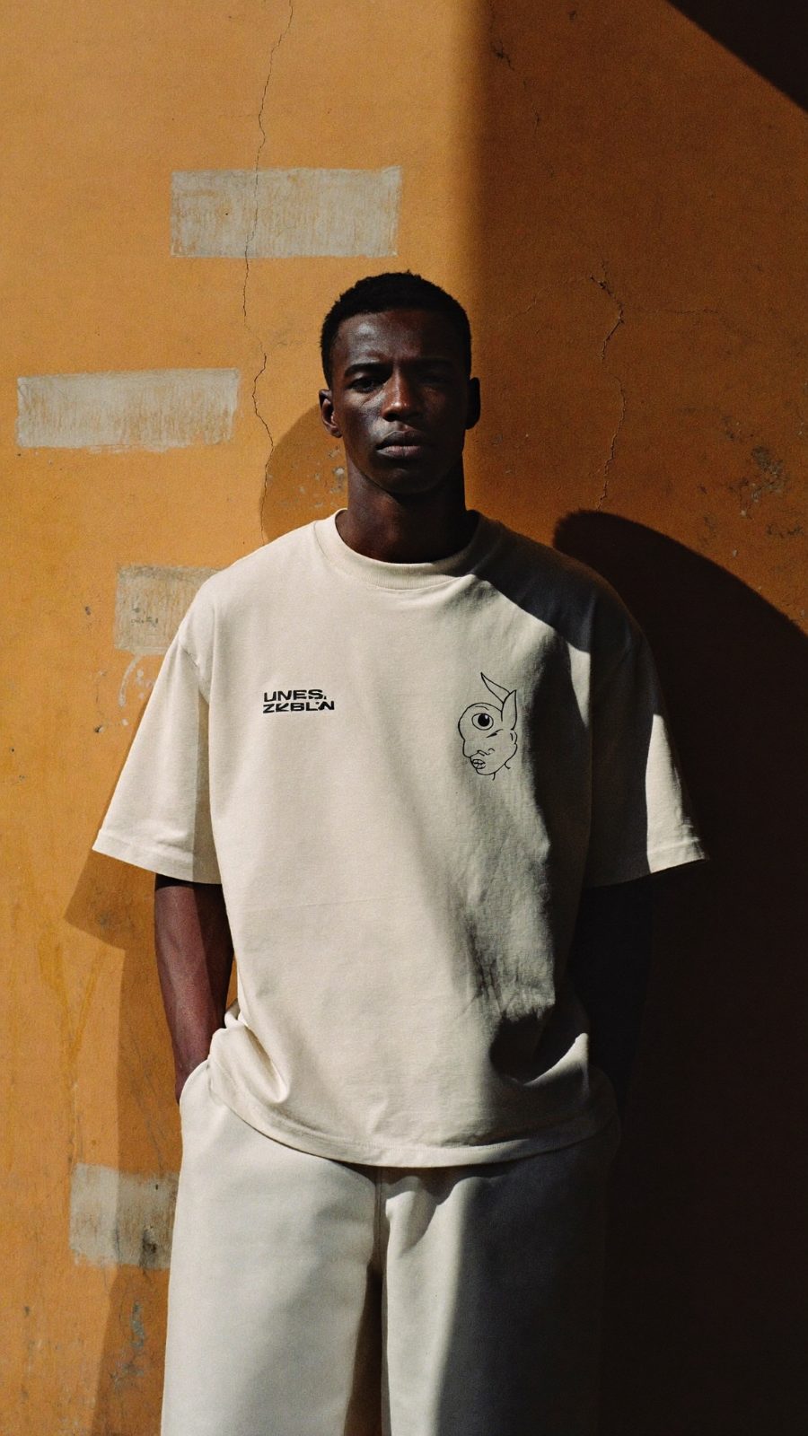 Model in bold streetwear against a painted concrete wall in hard midday sun.